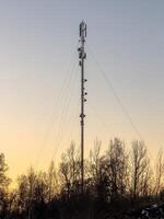 A tall communication tower stands silhouetted against a warm sunset sky, with sparse trees in the foreground. The tower is equipped with various antennas and equipment, emphasizing modern technology photo