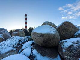 Osinovets Lighthouse rises in the background behind large snow-covered boulders, creating a dramatic contrast against the clear evening sky. The golden glow of the setting sun highlights the winter photo