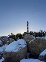 Osinovets Lighthouse rises in the background behind large snow-covered boulders, creating a dramatic contrast against the clear evening sky. The golden glow of the setting sun highlights the winter photo