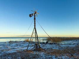 An old windmill with wooden blades stands on the snowy shore of Lake Ladoga, with clear skies and distant reeds lining the horizon. The tranquil winter landscape captures the beauty of the northern photo