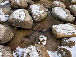 Large smooth stones partially covered in snow emerge from the frozen shoreline of Lake Ladoga. The icy water flows between the rocks, creating a striking contrast between the white frost and the dark photo