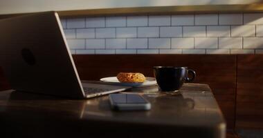 A small table in a cafe with an open laptop, a mobile phone, a donut and coffee standing on it, there are no people video