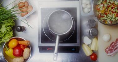 A small pot of boiling water is on the stove, vegetables and bacon are on the table next to it, top view, there are no people video