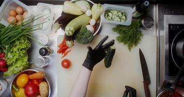 A woman in disposable black gloves cuts cucumbers standing near a table full of vegetables, close-up of her hands, top view video