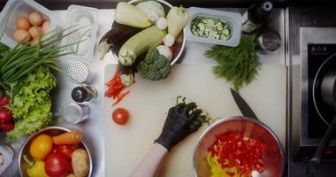 A woman in disposable black gloves adds a sliced cucumber to a salad bowl, standing near a table filled with vegetables, close-up of her hands, top view video