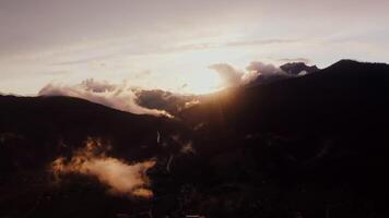 The light of the rising sun breaks through fluffy white clouds over the peaks of the Alps. Village in a lowland between mountains at dusk, drone , Austria video