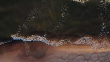 Top view from a drone of the coastline at high tide. Foamy sea waves crash against the shore, leaving patterns on the sand. Bird's-eye view. video