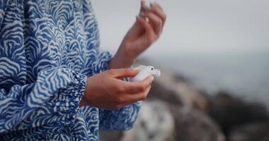 A beautiful young woman takes out wireless headphones from the box and puts them in her ears, sitting on the rocks on the seashore. 4k , red komodo video