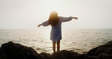 A young woman moves her arms smoothly, spread them to the sides, standing on a rocky seashore in the light of the setting sun. filming from the back. 4k , red komodo video