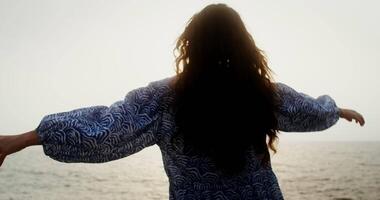 A woman smoothly moves her arms, spreading them to the sides, standing on the sea coast in the light of the setting sun and looking into the distance. filming from the back. 4k , red komodo video