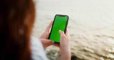 fechar-se do uma telefone com uma verde tela dentro a mãos do uma mulher digitando em uma Smartphone enquanto sentado em a pedras em a Beira Mar. croma chave, brincar, cópia de espaço. 4k , vermelho Komodo video