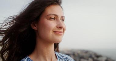 A beautiful young woman with flowing curly dark hair smiling looking at the sea, standing on a rocky coast, close-up of a smiling face from the side. 4k , red komodo video