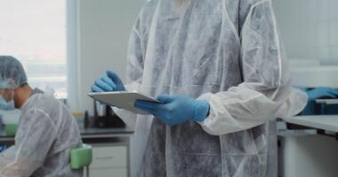 A male laboratory assistant in disposable medical clothing looks up from using a digital tablet and looks directly into the camera while working in a modern laboratory. 4k , red komodo video