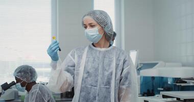 A young female laboratory assistant in disposable sterile clothes breaks away from examining a test tube with liquid and looks into the camera while working in a laboratory team. 4k , red komodo video