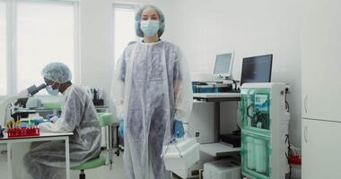 A female laboratory assistant in disposable clothes, in a medical mask, holds a box with test samples and looks into camera, standing in laboratory with working doctors. 4k , red komodo video