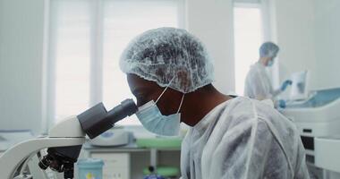 Close-up of the face of a laboratory assistant in a medical mask and disposable cap, looking through a microscope while checking blood, working as a team. 4k, red komodo video