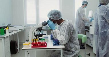 A male laboratory assistant of African American appearance, dressed in sterile clothes, looks through a microscope while checking a blood test in a modern laboratory. 4k, red komodo video