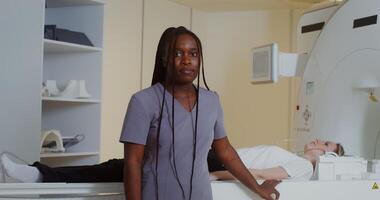 A young female doctor with long afro braids looks straight into the camera as she stands in front of a CT scan machine with a patient lying on it. in 4k, red komodo video