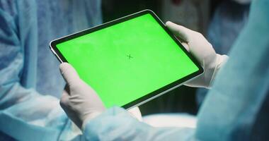 Close-up of a digital tablet with a green screen in the hands of a doctor in rubber gloves standing near a surgeon during an operation. Chroma key, mockup, copy space. in 4k, red komodo video
