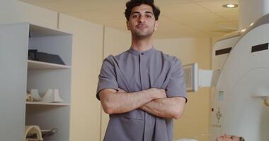 A young curly-haired doctor with stubble on his face looks straight into the camera with a smile and crosses his arms over his chest, standing in front of a CT scan machine. in 4k, red komodo video