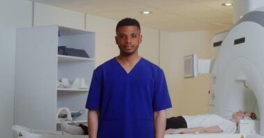 A young male doctor looks into the camera while standing in front of a CT scan machine with a female patient lying on it. in 4k, red komodo video