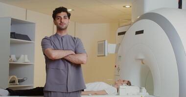 A curly-haired doctor with stubble on his face looks into the camera with his arms crossed over his chest, standing in front of a CT scan machine with a patient lying on it. in 4k, red komodo video
