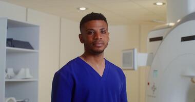 A young male doctor looks at the camera while standing in front of a CT scan machine in a modern medical center. in 4k, red komodo video