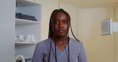 Young African American doctor with long afro braids looks directly into the camera while standing in front of a CT scan machine in a modern medical center. in 4k, red komodo video