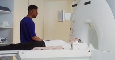 A young male doctor of African American appearance presses a button on the control panel of an MRI machine, starting a computed tomography of brain of a young female patient. in 4k, red komodo video