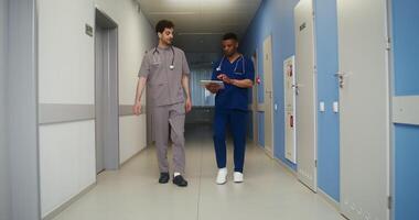 Two men dressed as medical personnel are smiling while looking at a tablet while walking in the corridor of the medical center. in 4k, red komodo video