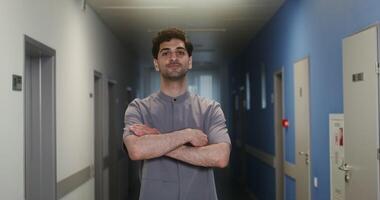 Young curly male doctor with stubble on his face crosses his arms over his chest and looks directly at the camera with a smile, standing in the corridor of the clinic. in 4k, red komodo video