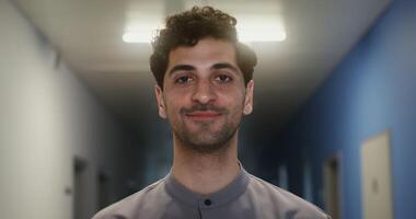 Young curly male doctor with stubble on his face smiling looking directly at the camera, standing in the corridor of the medical center, close-up. in 4k, red komodo video