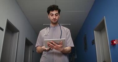 A young curly-haired male Caucasian doctor with a phonendoscope around his neck uses a tablet while walking along the corridor of the hospital. in 4k, red komodo video