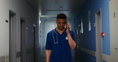 A male doctor with a phonendoscope around his neck smiles while talking on a mobile phone while walking along the corridor of the medical center. in 4k, red komodo video