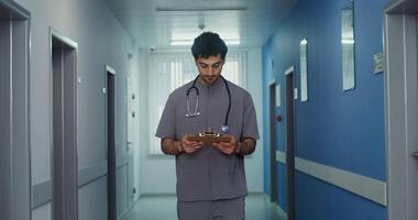 A male doctor with a phonendoscope around his neck walks along the bright corridor of a medical clinic and looks into a notebook that he holds in his hands. in 4k, red komodo video