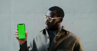 Young african american man smiling looking at camera while holding mobile phone with green screen, standing outdoors, copy space, mockup, chroma key. in 4k, red komodo video