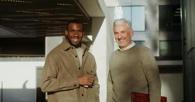 två män av annorlunda nationaliteter med en stor ålder skillnad är leende ser på de kamera medan stående nära de kontor Centrum under en kaffe ha sönder på en solig dag. i 4k, röd komodo video