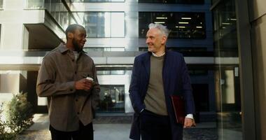An African-American man with a cup of coffee and a gray-haired Caucasian man with a laptop are smiling while talking in the business center of the city on a sunny day. in 4k, red komodo video
