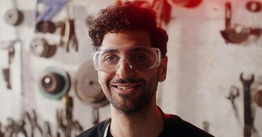 A young man in goggles smiles looking directly at the camera, against the background of a wall with work tools. Close-up of a man's face. in 4k, red komodo video