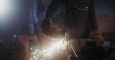 A blacksmith of African American appearance in goggles and an apron cuts off a piece of metal workpiece using an angle grinder. Close-up, motion from the man's hands to his face. 4k, red komodo video