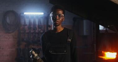 African-American man in goggles and work clothes looks at the camera with a serious look while standing in a modern forge with angle grinder in his hands. Zooming to man's face. 4k, red komodo video