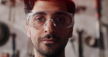 A close-up of the face of a young man in protective glasses who looks up from a digital tablet and looks up at the camera with a smile. in 4k, red komodo video
