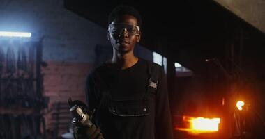 Young African-American man in goggles and work clothes smiling looking at the camera while standing in a modern forge with an angle grinder in his hands. 4k, red komodo video