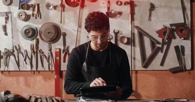 A young blacksmith in working clothes smiling looking at the camera distracted from work at the tablet, sitting at the work table in the forge. in 4k, red komodo video