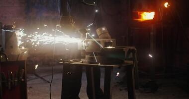 A young blacksmith of African American appearance in goggles and work clothes uses an angle grinder while working in a modern forge. Metal sparks fly in all directions. 4k, red komodo video