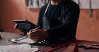 un' giovane maniscalco vestito per opera usi un' tavoletta per creare un' schizzo di un' futuro Prodotto, seduta a il suo scrivania nel un' fucina pieno di Lavorando Strumenti. nel 4k, rosso komodo video