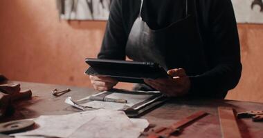 A blacksmith in working clothes smiling looking at camera distracted from work at tablet, sitting at the desk in the forge. Close-up, moving from the men's hands to face. in 4k, red komodo video