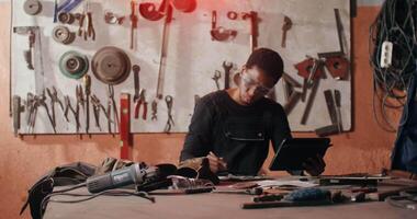 African-American man in goggles and work clothes uses a digital tablet while creating a sketch for a future product, sitting at work table in a forge among a variety of tools. in 4k, red komodo video