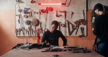 Two blacksmiths in work clothes are drawing a sketch of a future product, working together and using a tablet to search for ideas, in a workshop full of working tools. in 4k, red komodo video