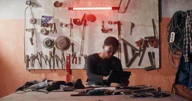 An African-American man in goggles and work clothes is sketching a product using a digital tablet while sitting at a desk in a forge among many tools. in 4k, red komodo video